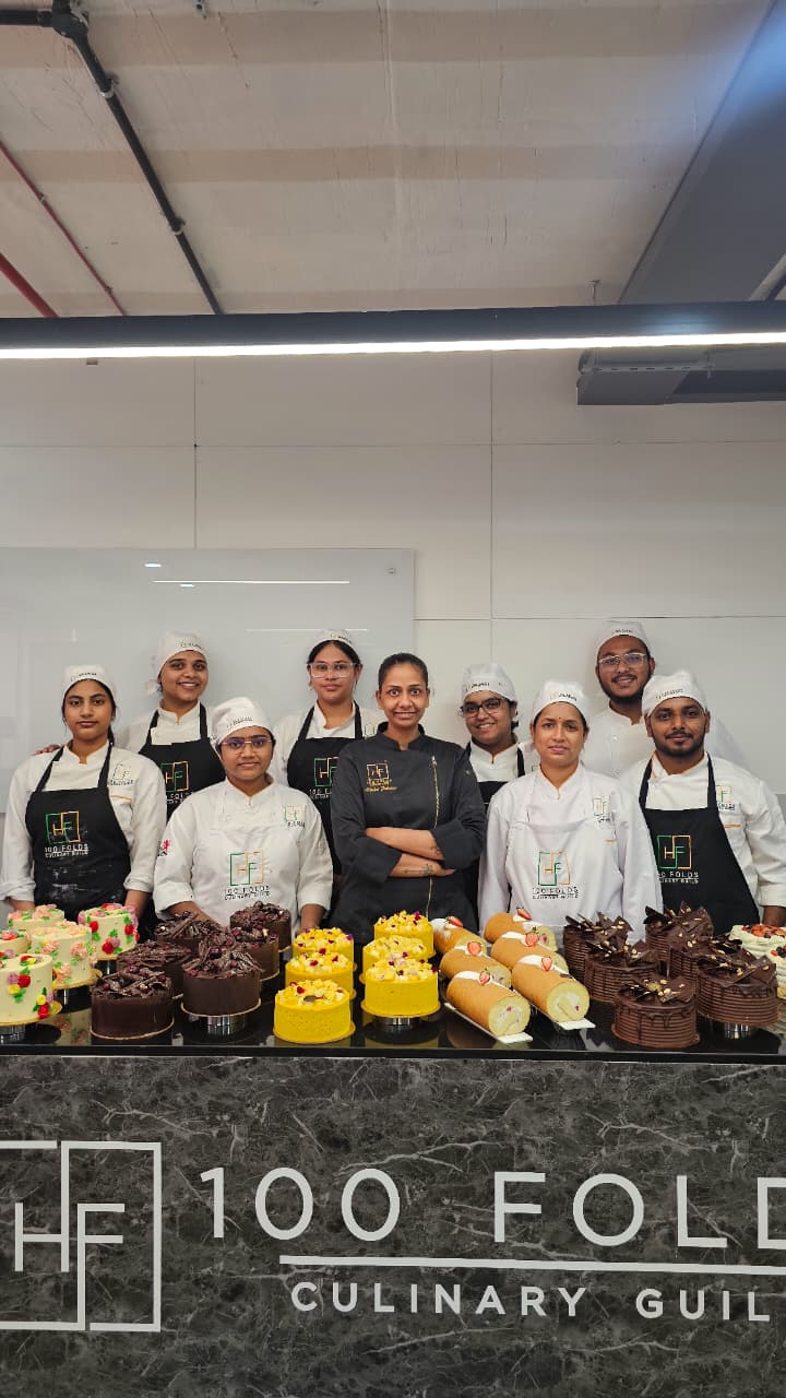 Students and instructor at 100 Folds Culinary Guild Hyderabad showcasing handmade chocolate, mango, and fresh cream cakes during hands-on baking class.