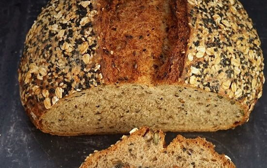 A rustic whole wheat bread loaf with a sliced crust, topped with oats and seeds, with several slices cut and stacked in front, set on a dark slate surface.