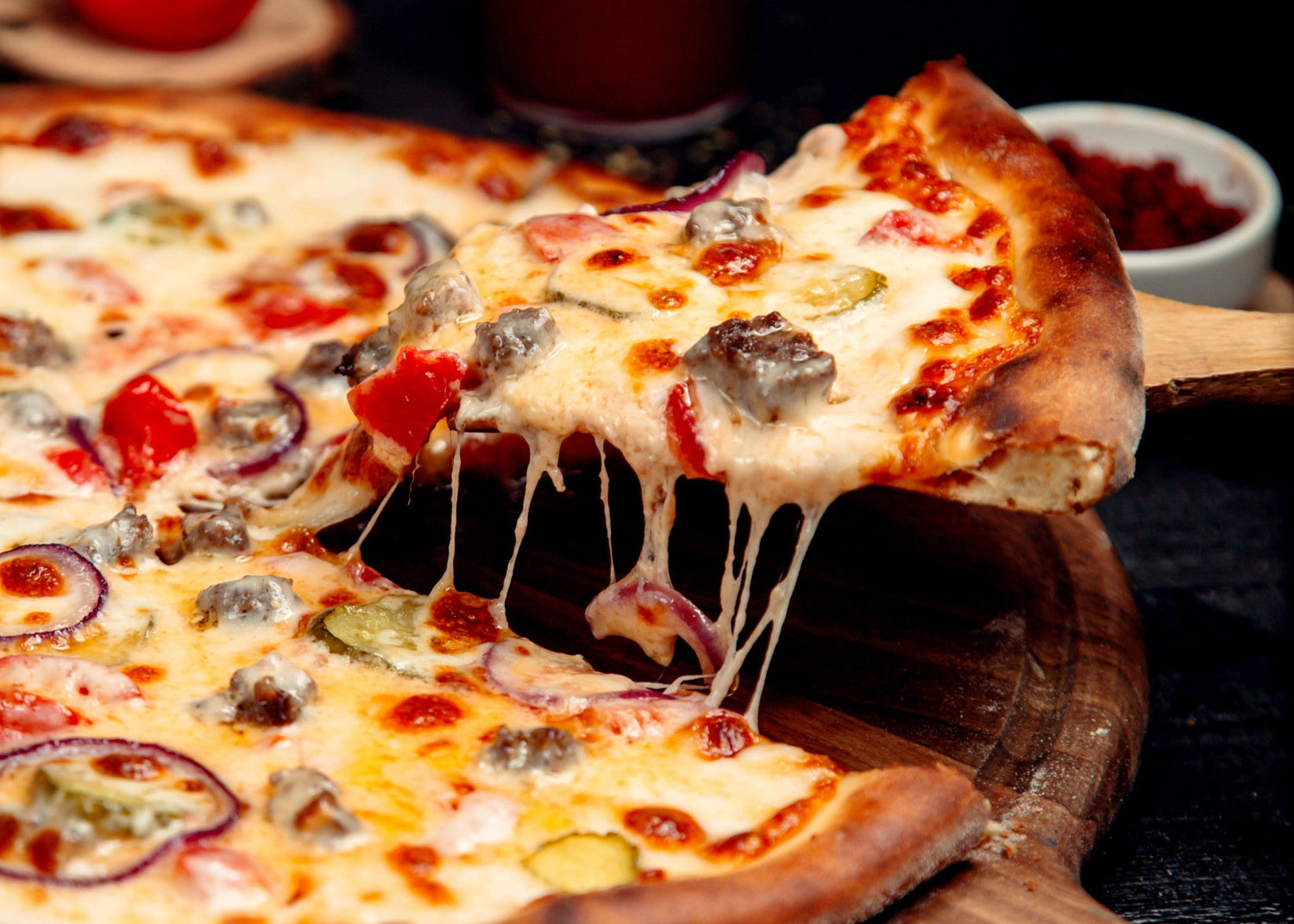 Hot pizza with a slice being lifted, showing melted cheese, on a wooden board with a blurred background.