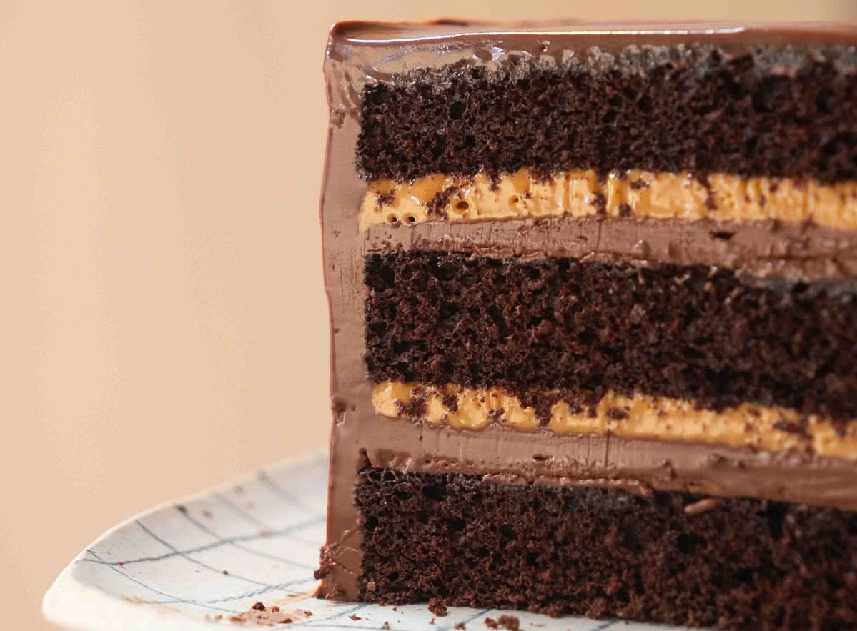 A close-up of a layered chocolate cake slice on a white plate with a subtle grid pattern, set against a soft beige background.