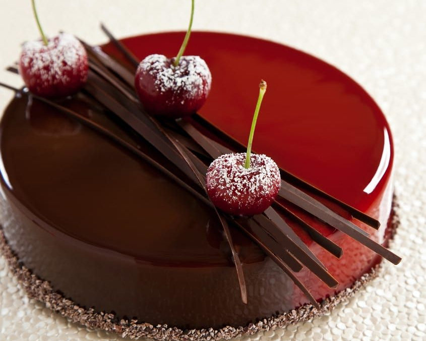 A round chocolate cake with a glossy dark brown chocolate glaze and a contrasting glossy red upper section, decorated with three cherries dusted in powdered sugar and several chocolate shards on top, set on a textured cream background.