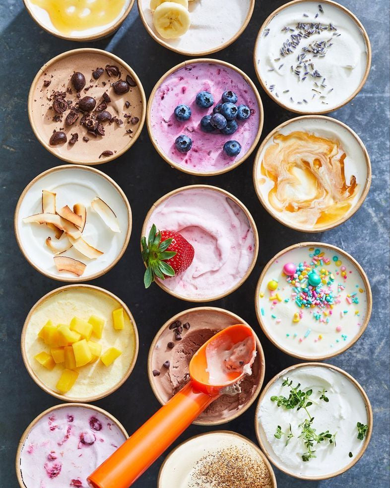 An overhead flat-lay of multiple small bowls filled with different yogurt or cream-based desserts, each topped with varied ingredients like blueberries, chocolate, fruit, sprinkles, and herbs. An orange spoon rests across the bowls on a dark textured surface.