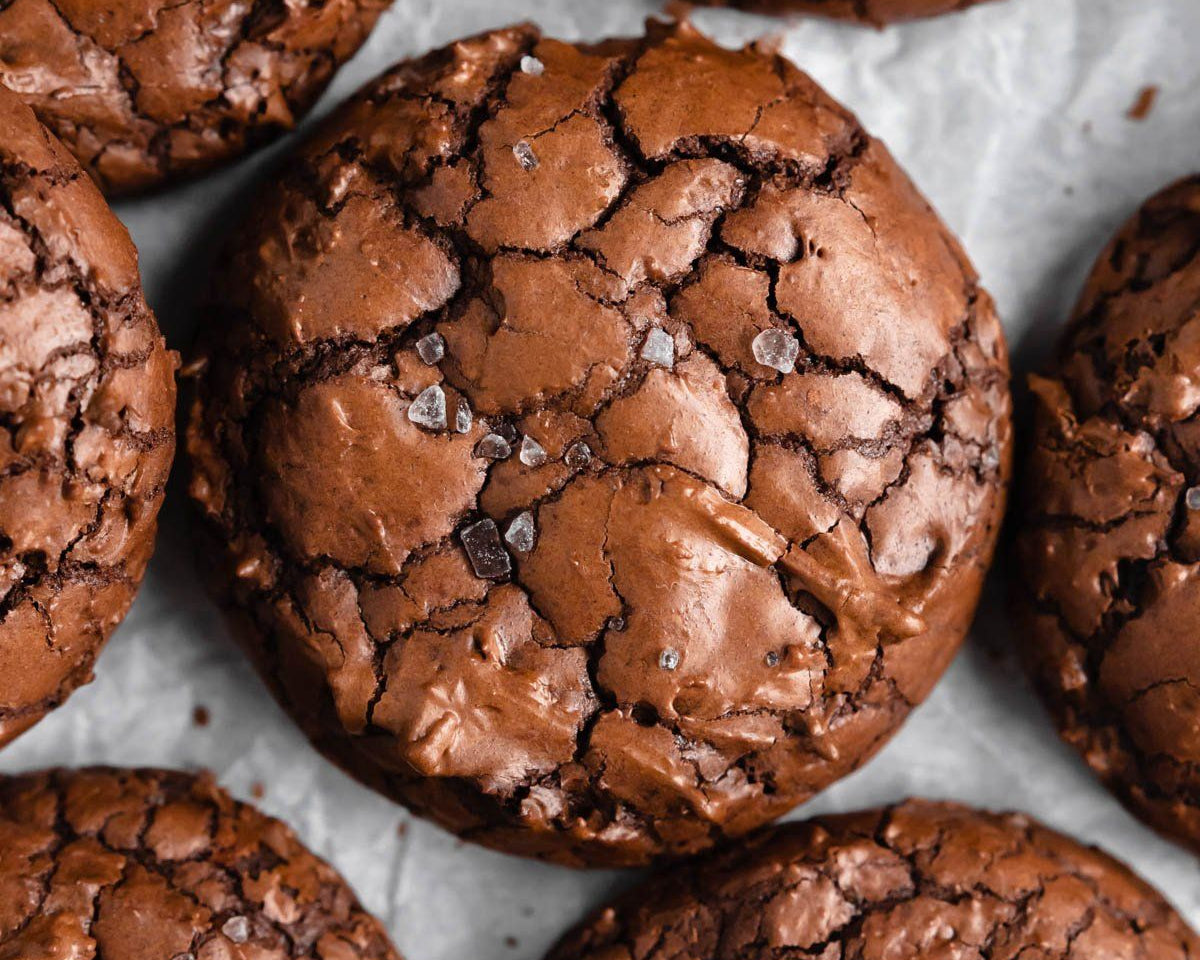 Close-up overhead view of several chocolate brownie cookies with a glossy, cracked surface and a few coarse salt crystals, arranged on a light gray surface.