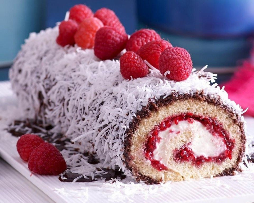 A raspberry and cream swiss roll cake, coated in white chocolate and shredded coconut, topped with fresh raspberries, served on a white plate.