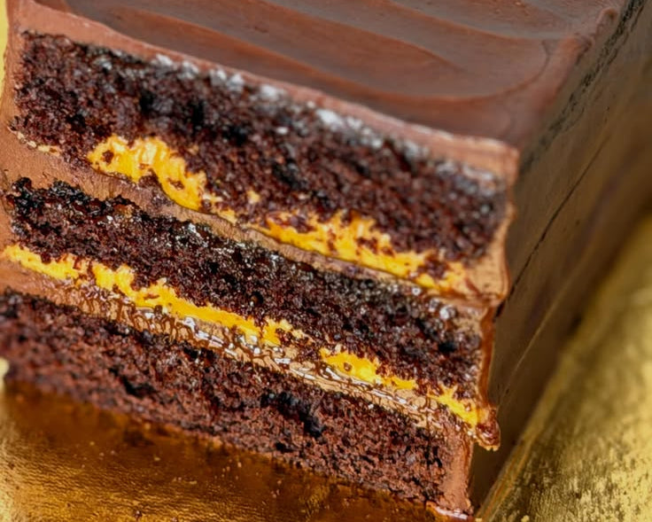 A close-up of a rectangular chocolate cake with two layers of dark chocolate sponge and a bright orange-yellow custard or caramel-like filling, topped with smooth chocolate frosting on a gold surface.